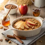 A close-up of a baked apple oatmeal dish in a white oval baking dish, topped with fresh apple slices and nuts. A jar of honey, an apple, and cinnamon sticks are on the wooden table.