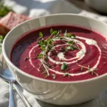 Bowl of creamy beet soup garnished with herbs and cream swirl