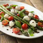 Roasted asparagus topped with cherry tomatoes, mozzarella, and fresh basil on a white plate.